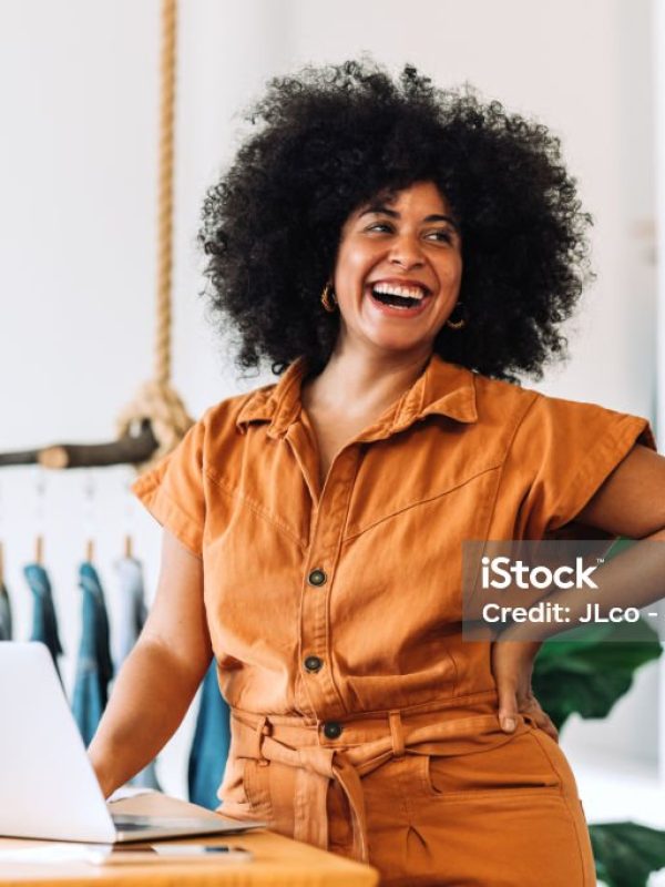 Ethnic small business owner smiling cheerfully while standing in her shop. Happy businesswoman managing her clothing orders on a laptop. Black female entrepreneur running an online clothing store.